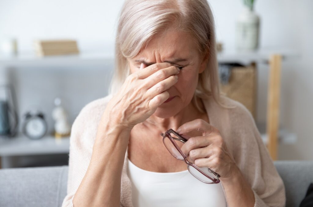 An older sitting on a couch holding a pair of glasses in their left hand and rubbing their eyes with their right hand due to blurry vision from cataracts.