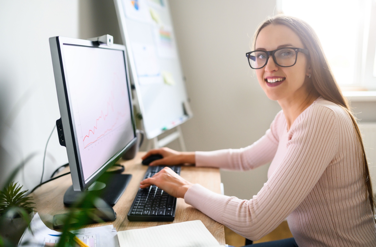 A smiling person wearing glasses faces the camera while sitting at a desk in front of a computer in a brightly lit room.