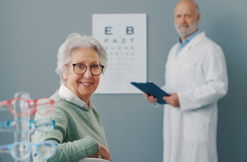 Senior woman with glasses looking behind her chair at the camera. In the background, a man in a white coat holding a clipboard stands beside a chart of letters.