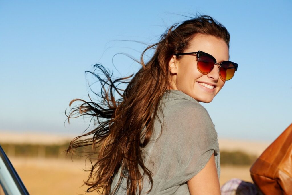 A smiling young woman with long brown hair blowing in the wind, wearing stylish tinted sunglasses while looking back over her shoulder outdoors.